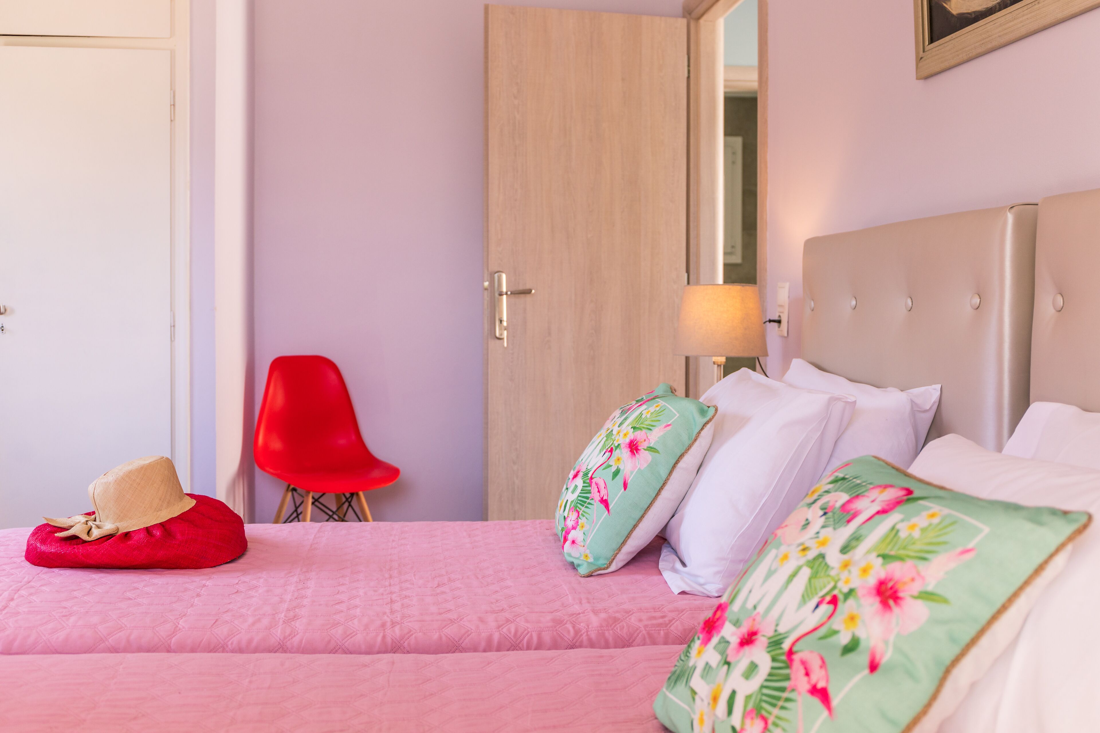 A hotel room with a bed, a red chair, a door, and a nightstand with a lamp.