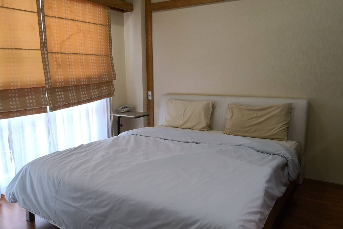 A neatly made bed with a white comforter and two pillows, a bedside table with a phone, and a window with a brown blind.