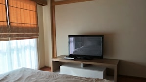 A hotel room with a bed, a television on a stand, and a window with blinds. - Choke-Set-Thee House (Ayutthaya)