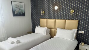Twee enkele bedden met witte lakens en handdoeken, een gouden hoofdbord, een wandspiegel en een klein nachtkastje met een lamp.