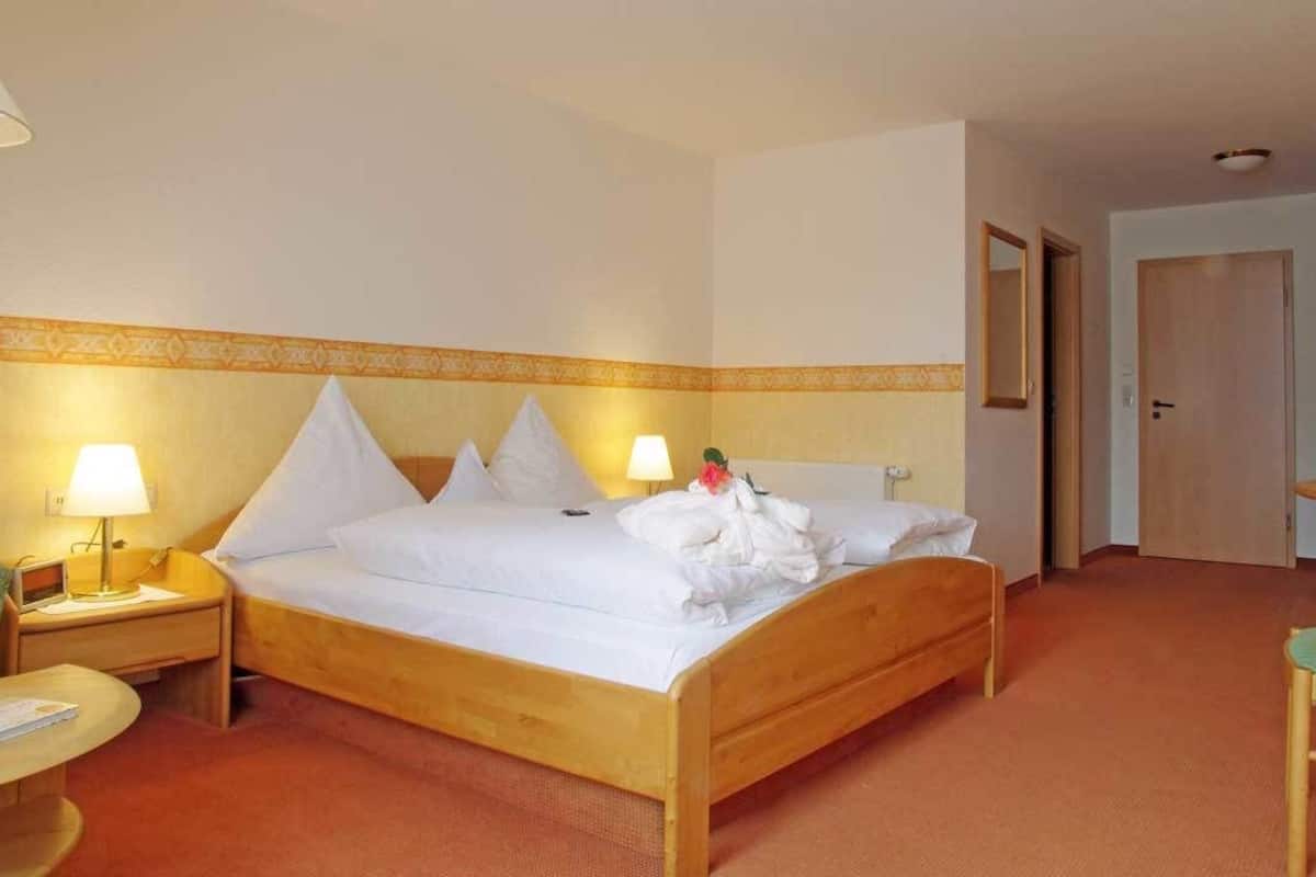 A hotel room with a bed, two bedside tables with lamps, a green chair, a small table with a book, and a television on a wooden stand.