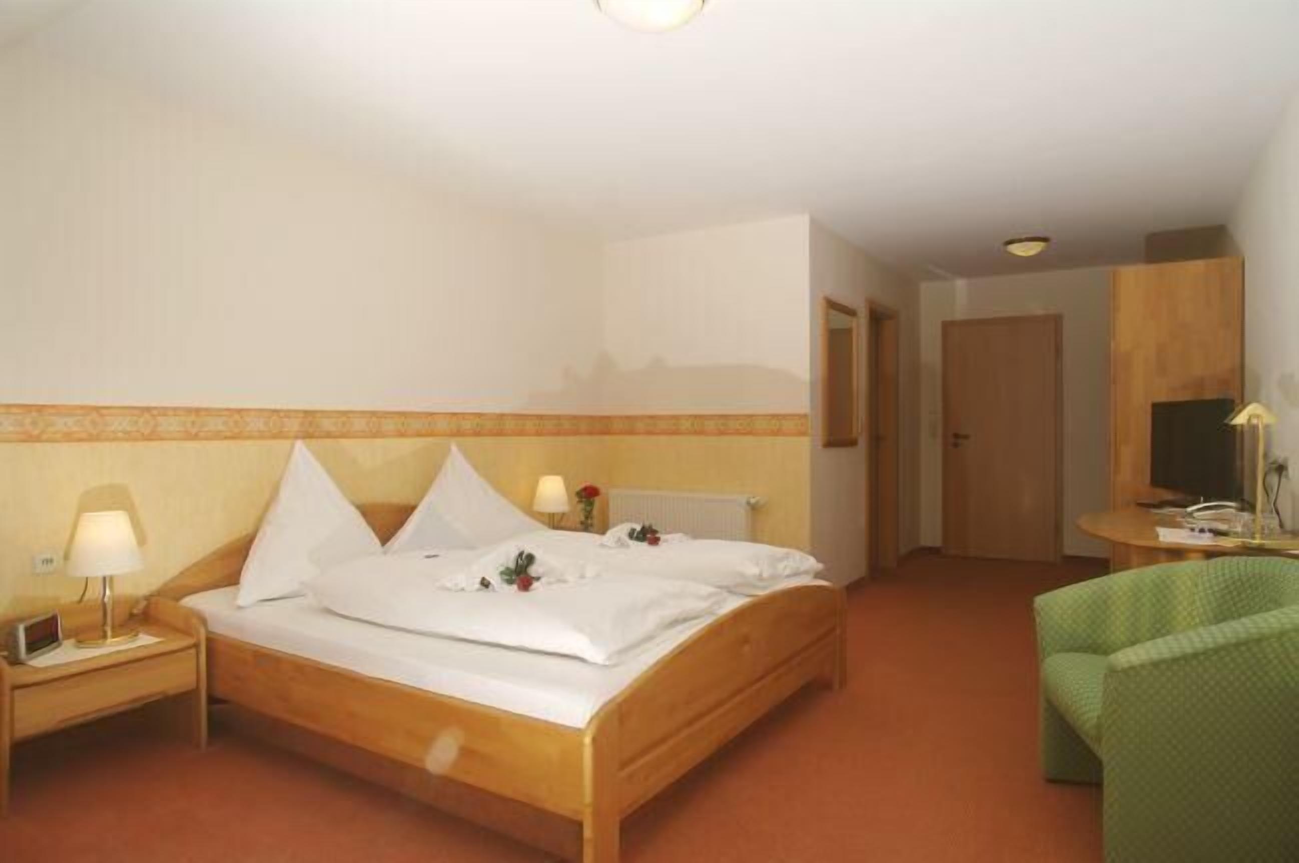 A hotel room with a wooden bed, a nightstand with a lamp, a green chair, and a small table with a lamp.