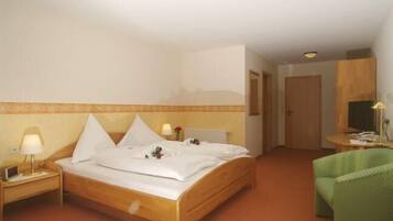 Habitación de hotel con cama de madera, mesita de noche con lámpara, una silla verde y una pequeña mesa con lámpara.