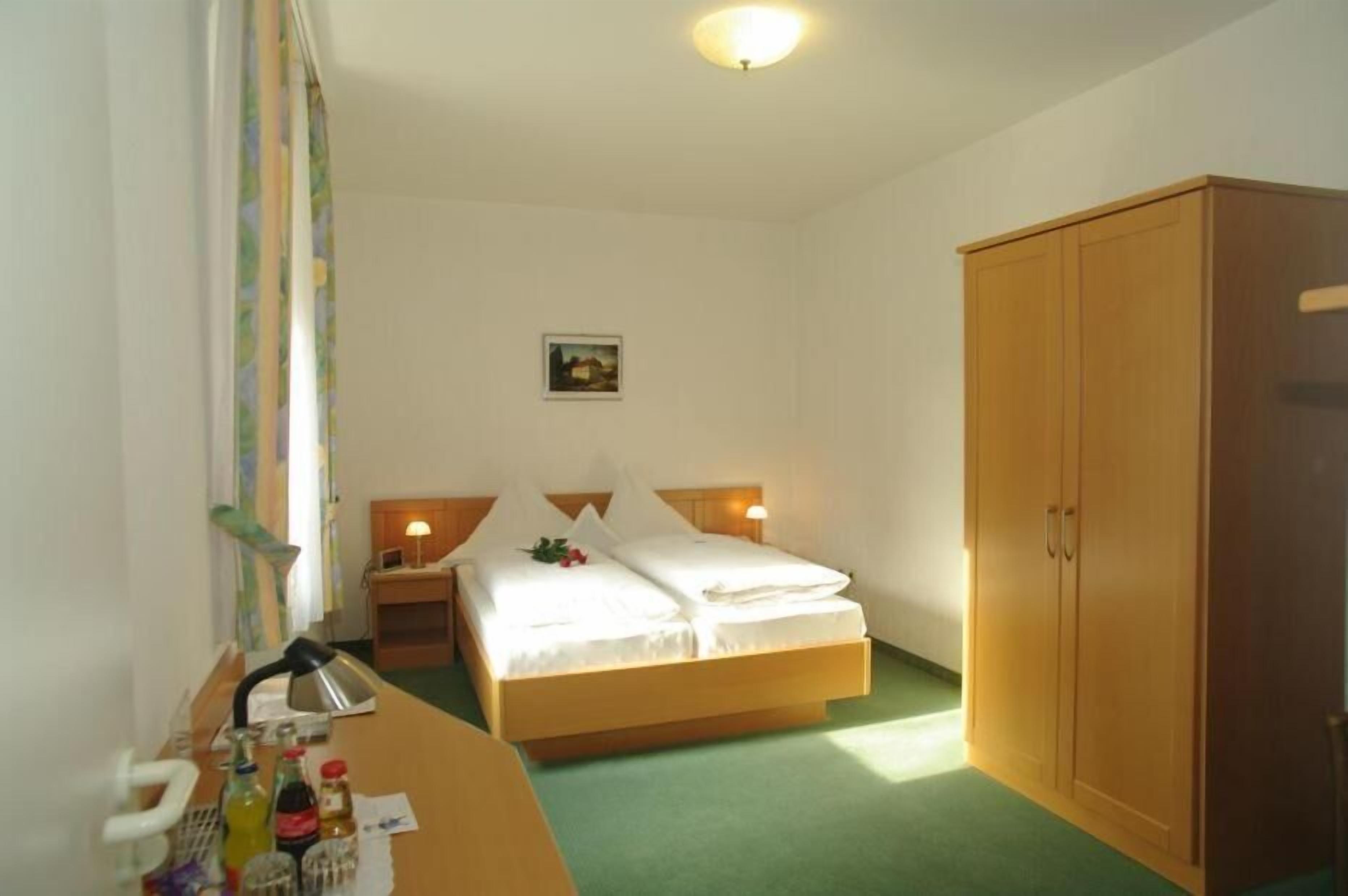 A single bed with a wooden headboard, a bedside table with a lamp, a wooden wardrobe, and a small framed picture on the wall.
