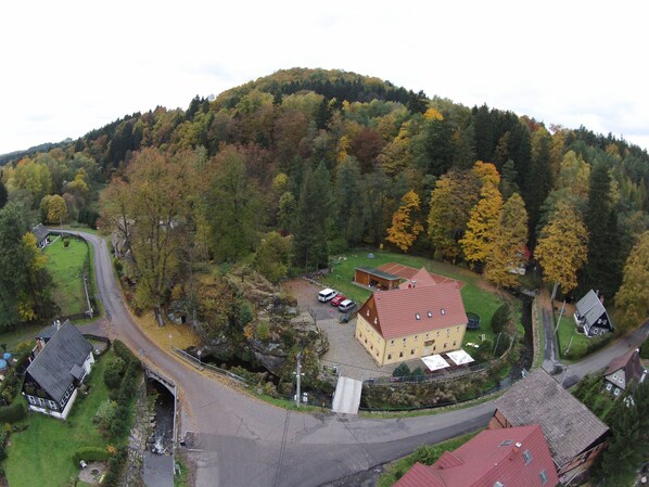 Vista aérea de uma área rural com um edifício amarelo imponente, cercado por árvores e uma estrada sinuosa.
