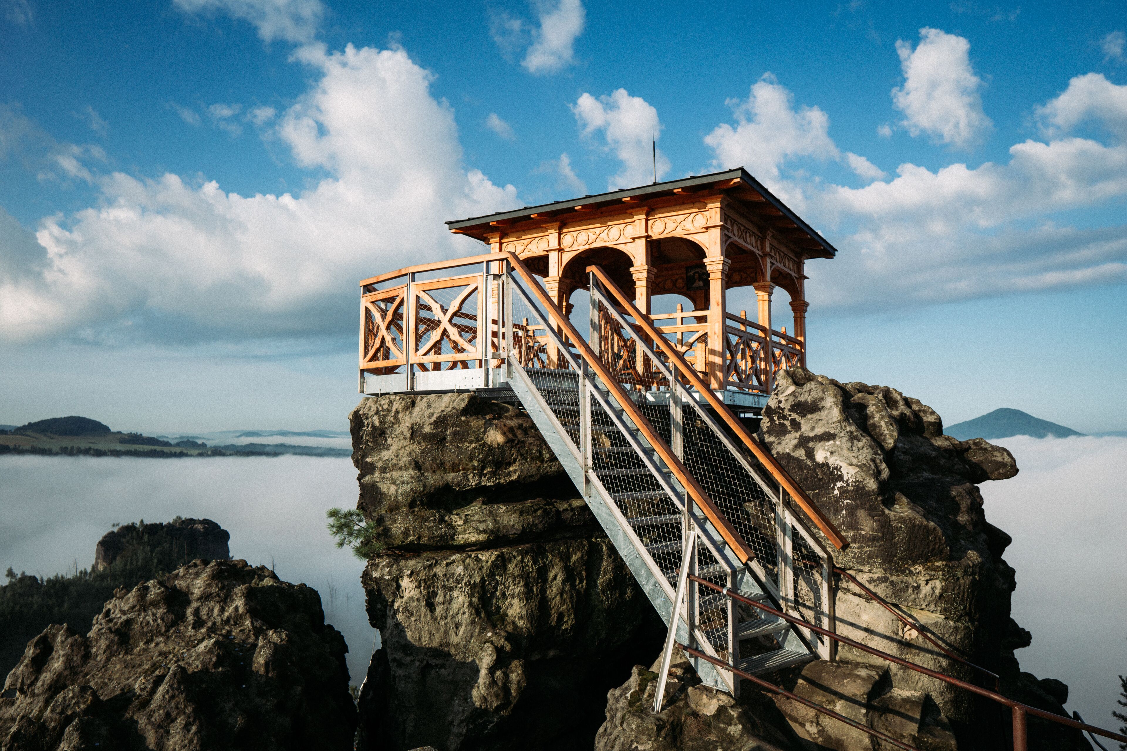 Una torre de observación de madera con una escalera sobre un afloramiento rocoso con vista a un cuerpo de agua y montañas distantes.