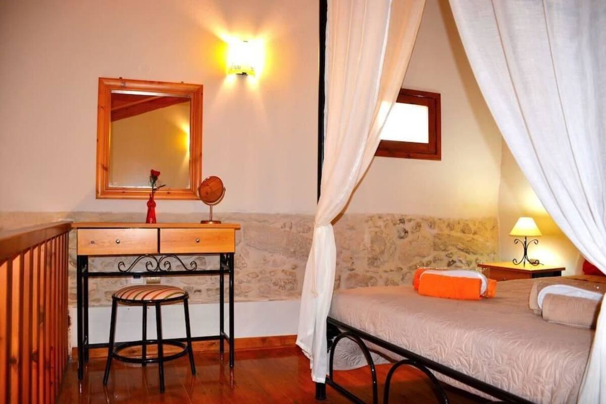 A bedroom with a bed, a nightstand, a lamp, a mirror, a wooden desk, and a chair.