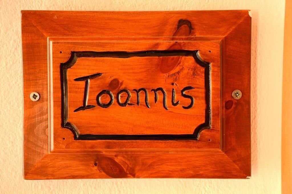 Une plaque nominative en bois portant le nom « Ioannis », fixée à un mur.
