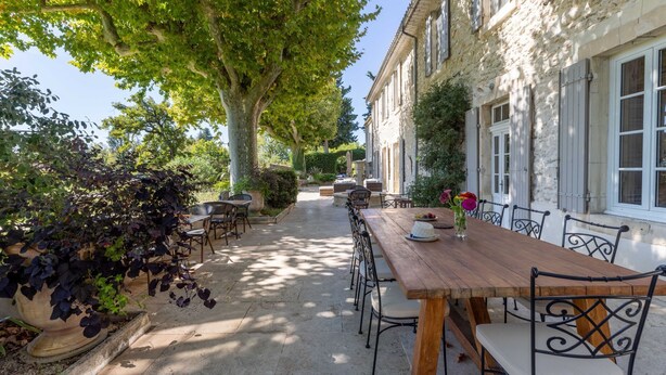 A courtyard with a wooden table and chairs, surrounded by stone buildings with white shutters.