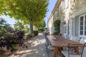 Une cour avec une table et des chaises en bois, entourée de bâtiments en pierre aux volets blancs.