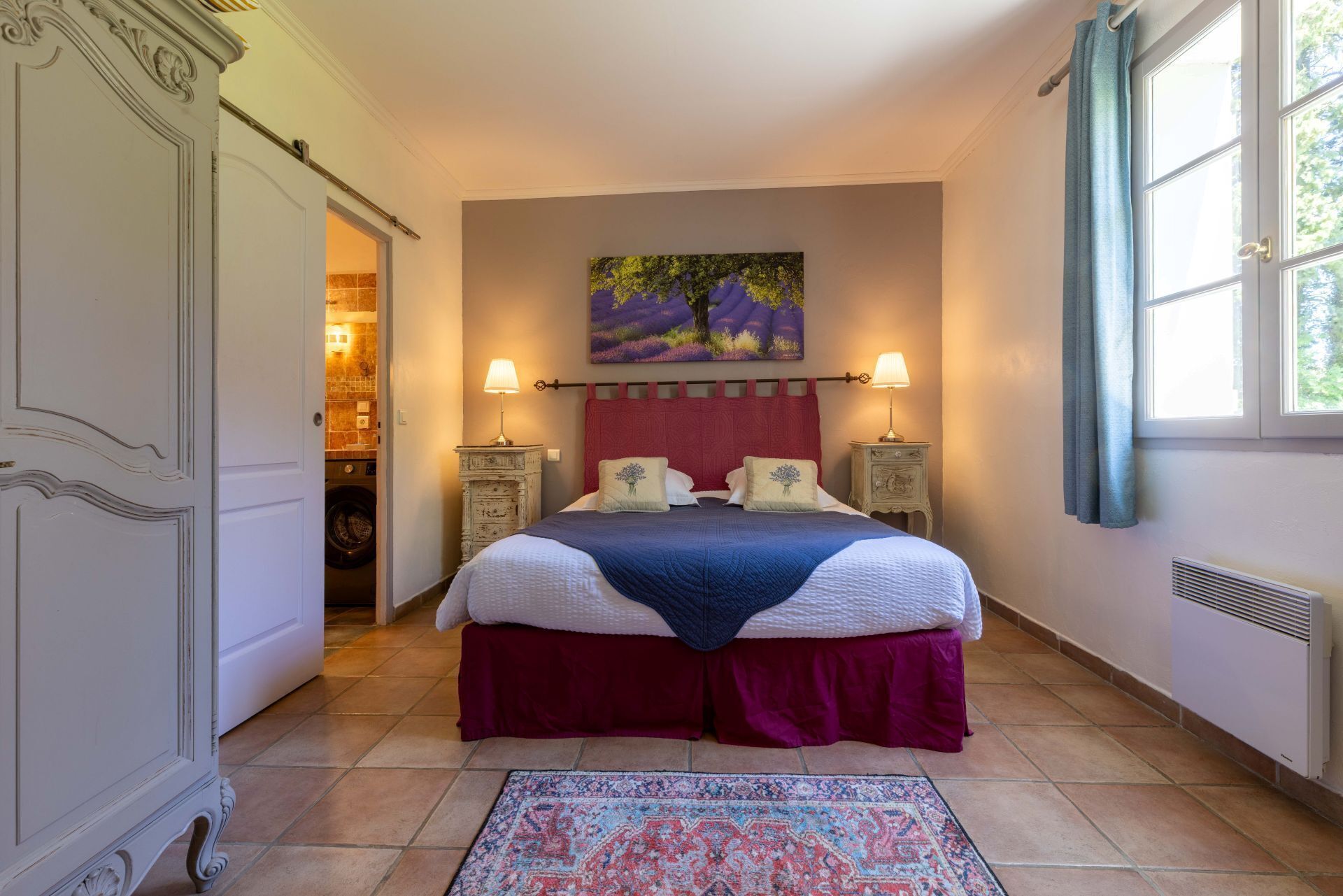 A bedroom with a large bed, two bedside tables, a painting on the wall, and a window with curtains.