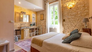 A bedroom with a stone wall, a large window, a bed with pillows, a nightstand, and a bedside lamp.