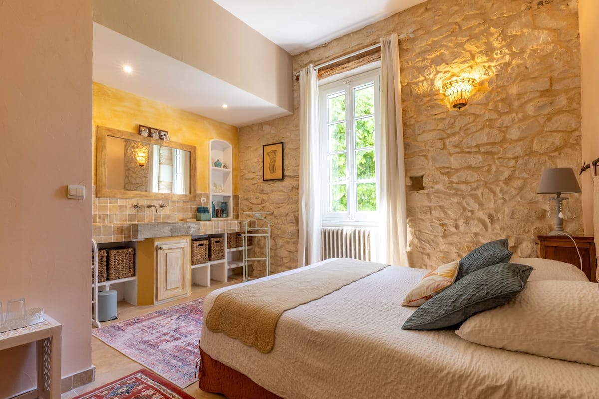 Ein Schlafzimmer mit Steinwand, großem Fenster, Bett mit Kissen, Nachttisch und Nachttischlampe.