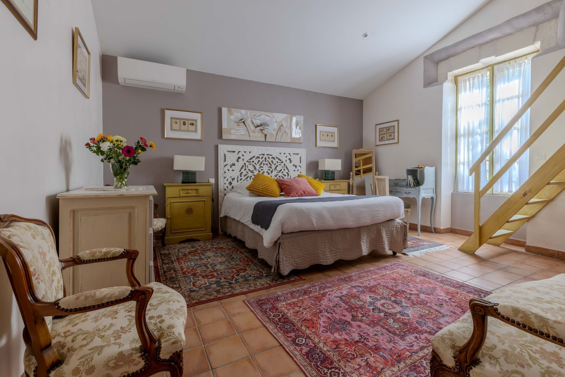 Quarto com cama, duas cadeiras, mesa de cabeceira, cômoda, janela com cortinas e escada.