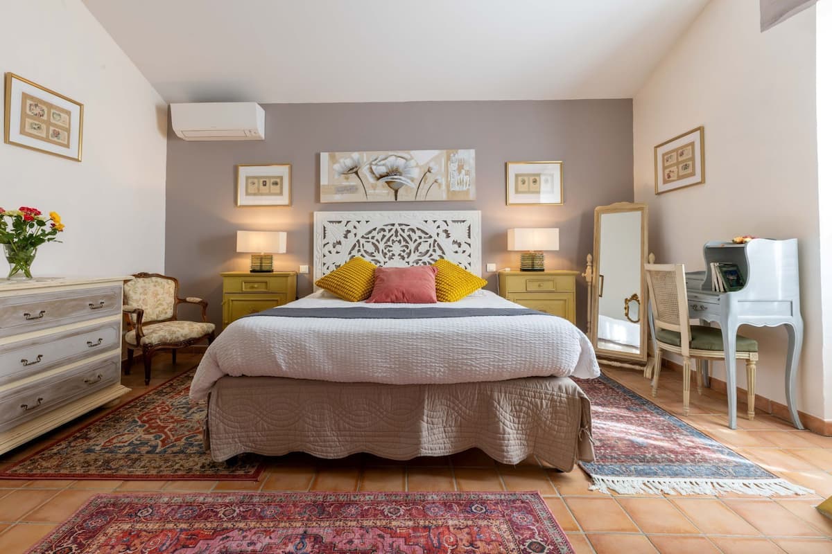 Ein Schlafzimmer mit einem großen Bett, zwei Nachttischen mit Lampen, einem Schminktisch, einem Stuhl, einem Schreibtisch, einem Spiegel und Bildern an den Wänden.