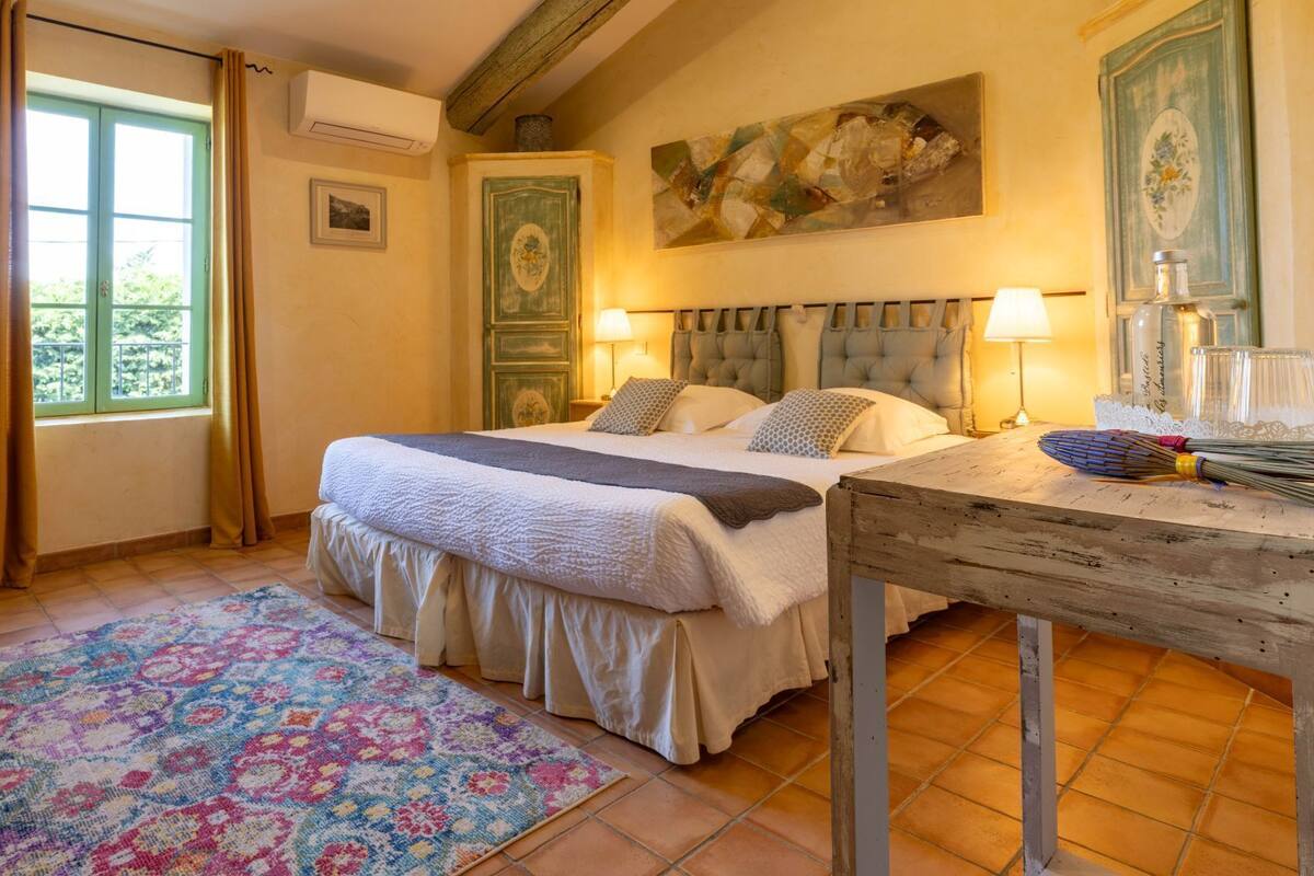 Ein Schlafzimmer mit Bett, Nachttischlampen, einem Holznachttisch, einer Heizung, einem Fenster mit Vorhängen und einem farbenfrohen Teppich.