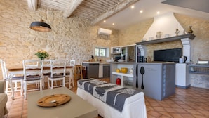 A cozy kitchen with a stone wall, a dining area with white chairs, and a bench with a blanket.