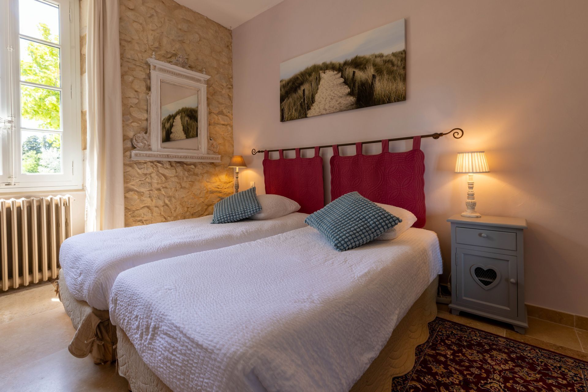 A bedroom with two beds, a stone wall, a window, a painting, a nightstand, and a radiator.