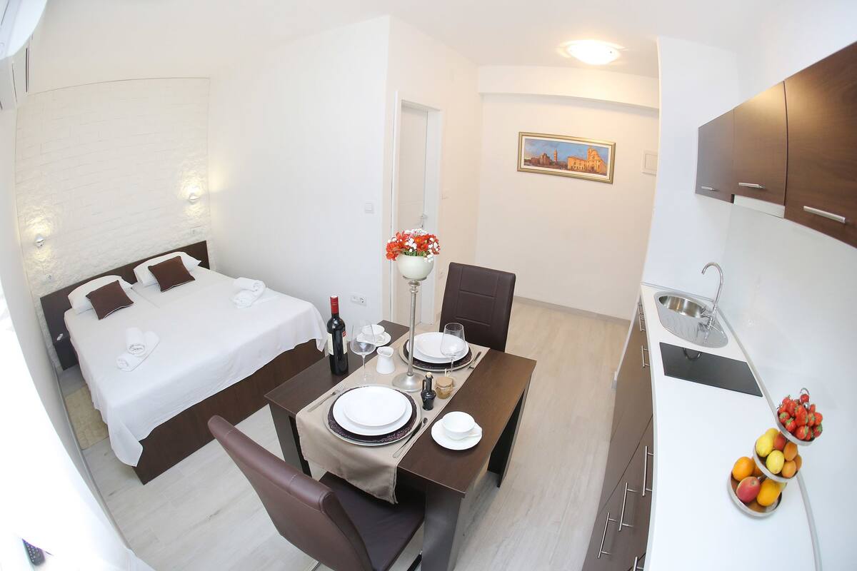 A hotel room with a bed, a dining table set for two, a small kitchen area, and a fruit bowl on the counter.