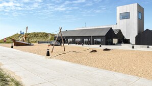 Edifici modern amb una gran façana de vidre, estructura de formigó i zona de platja amb un model de vaixell de fusta i un tobogan.