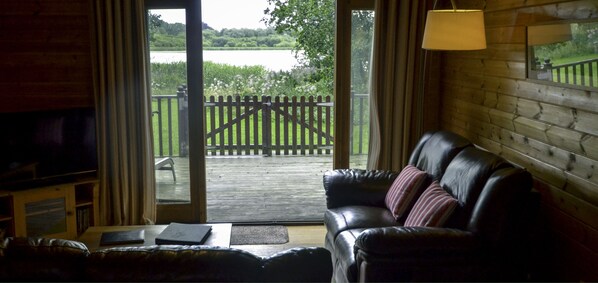 A cozy living room with a leather sofa, a television, and a wooden deck leading to a fenced garden. - The Tranquil Otter (Carlisle)