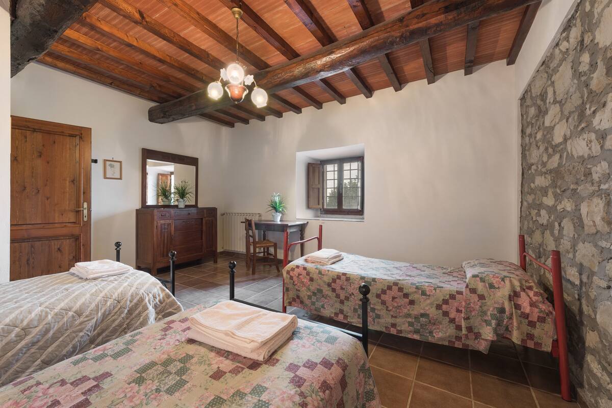 Una habitación con dos camas, vigas de madera en el techo, un escritorio con una silla, una ventana y una pared de piedra.