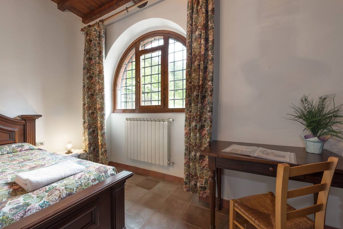 Un dormitorio con cama, escritorio, silla, radiador y ventana con cortinas.