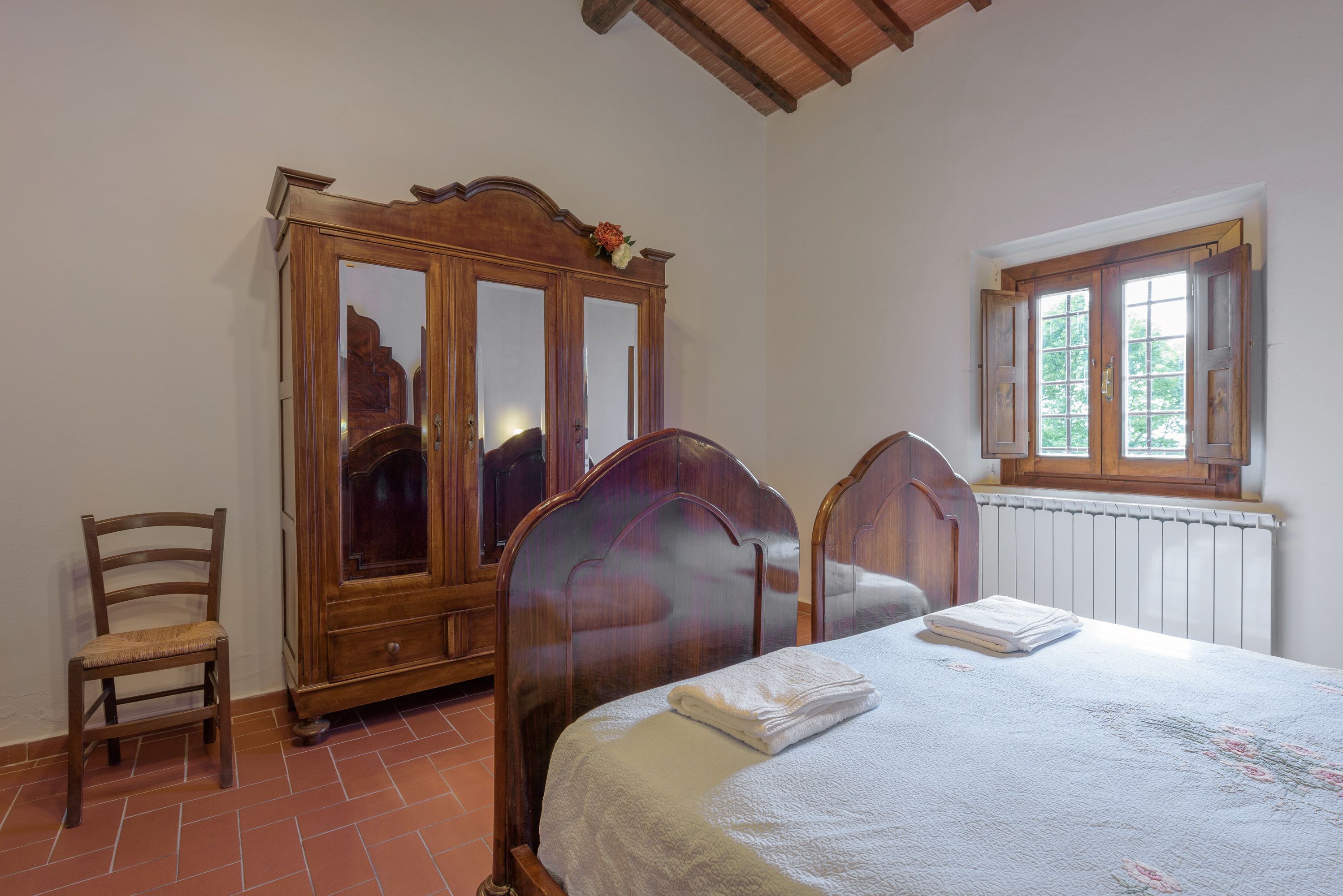 A bedroom with a wooden wardrobe, a single bed with a white blanket, a chair, and a window with a view of trees.