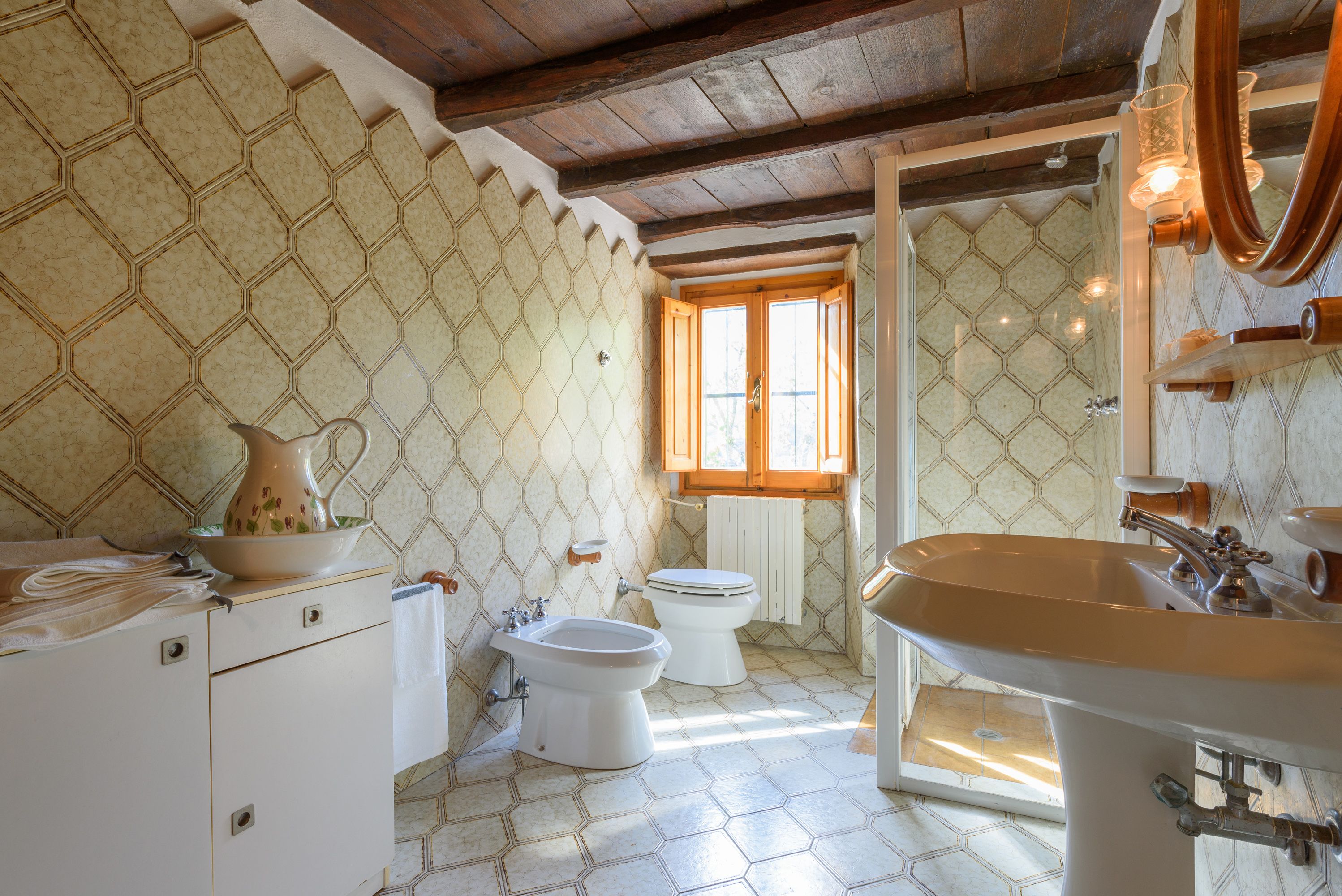 Une salle de bain avec un mur orné d’un motif hexagonal, un plafond en bois, une fenêtre, un lavabo, un bidet et des toilettes.