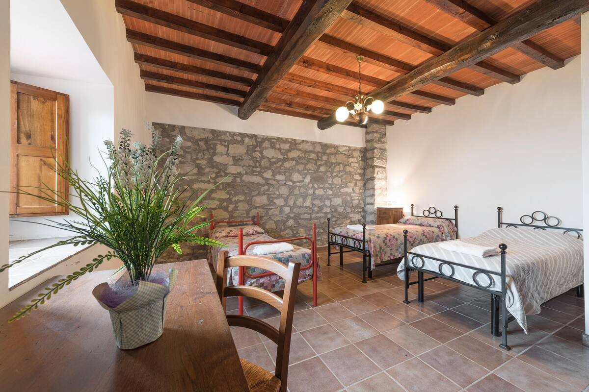 Un dormitorio rústico con pared de piedra, vigas de madera en el techo, una mesa de madera con sillas y dos camas de metal.