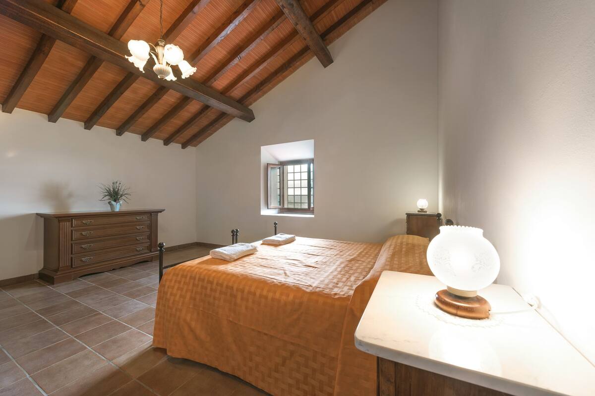 Un dormitorio con techo de madera, una cama con colcha estampada, un aparador, una mesita de noche y una lámpara de noche.