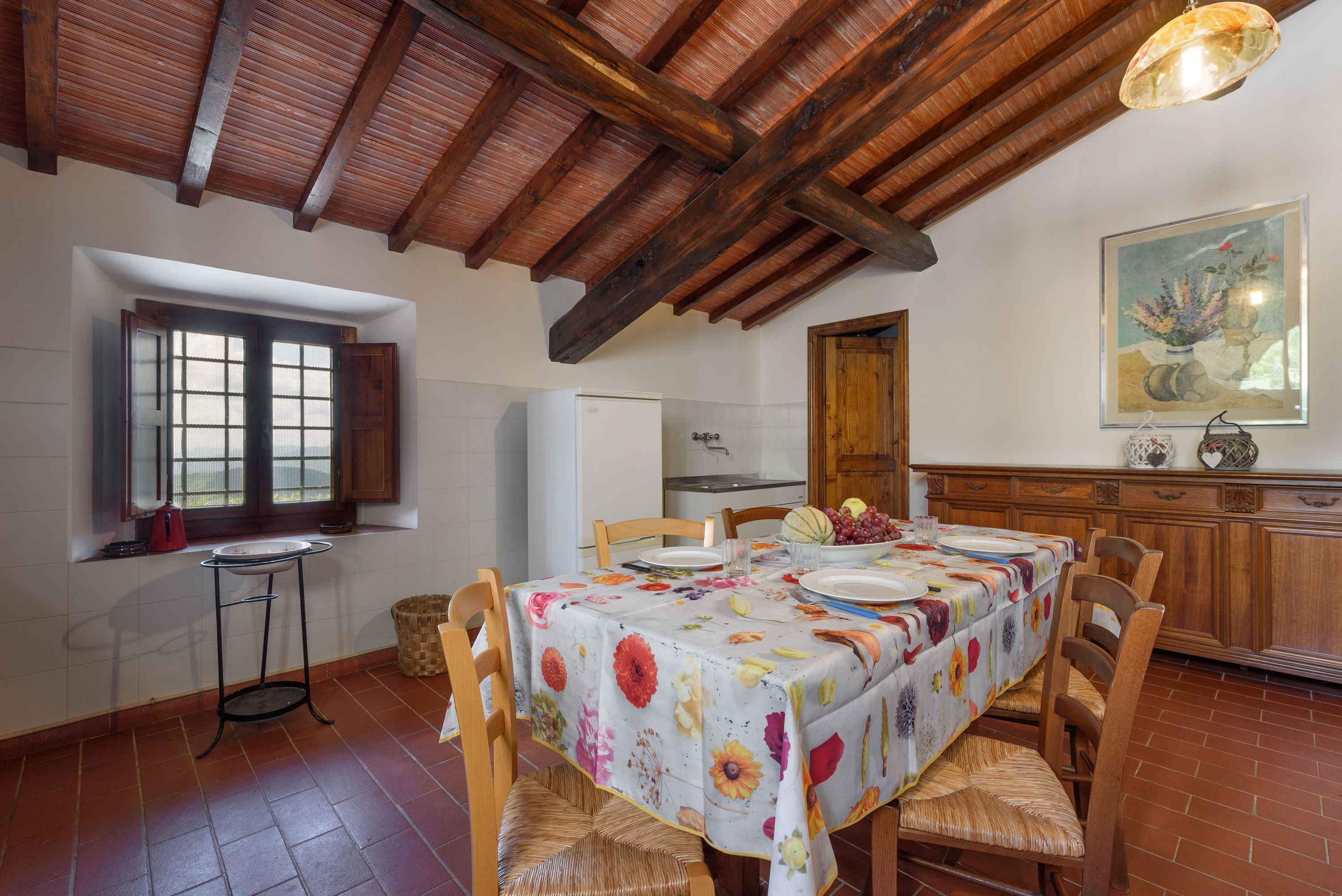 Un coin repas avec une table dressée pour quatre personnes, des chaises en bois, une fenêtre avec des volets, une cuisine en arrière-plan et un plafond en bois.