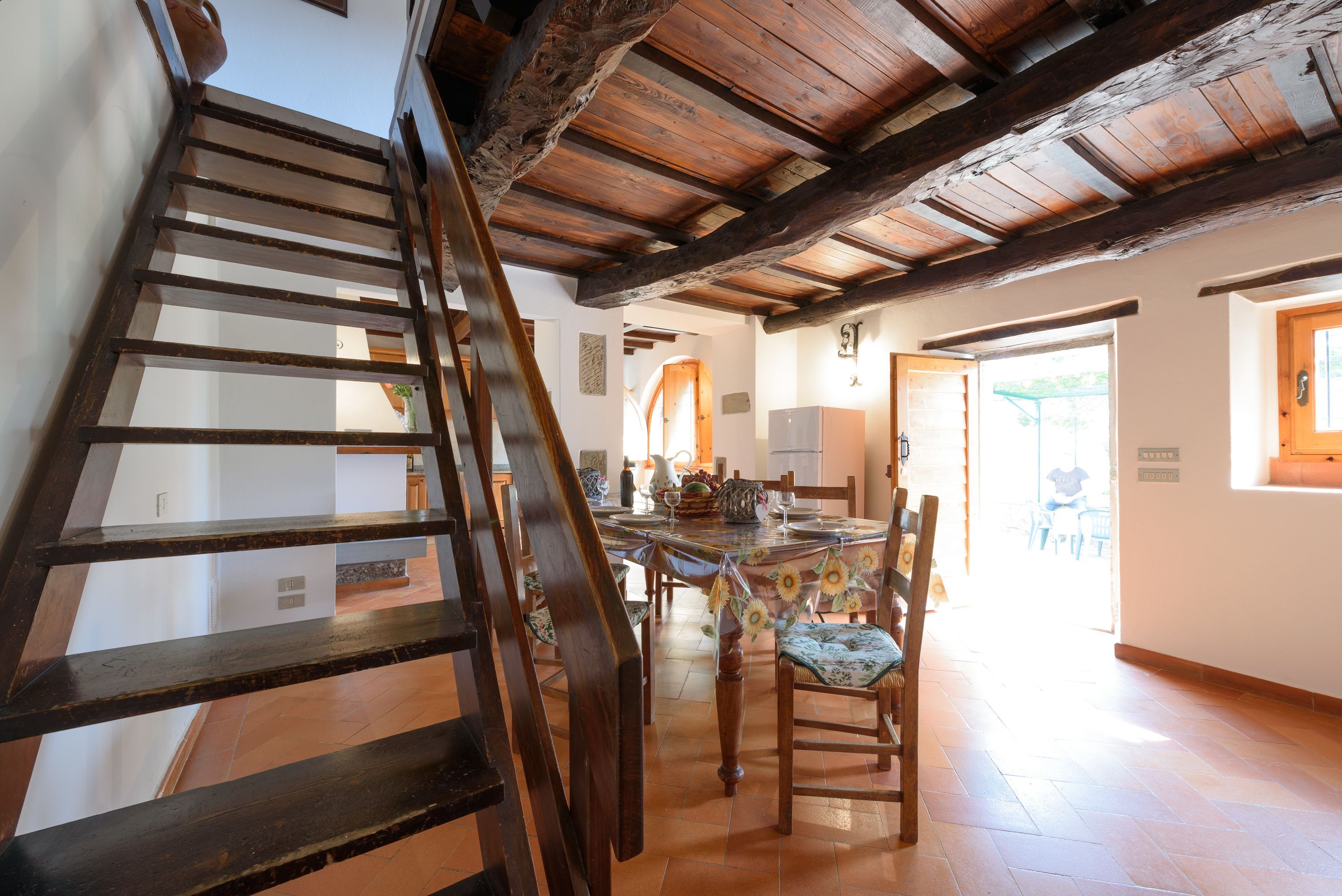 A staircase leading to a dining area with wooden chairs and a table, a kitchen area with a refrigerator, and a view of the outdoors.