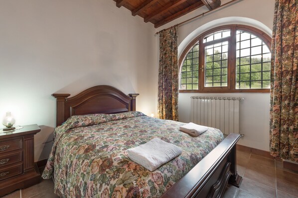 A bedroom with a wooden bed, floral bedspread, a nightstand, and a window with curtains.