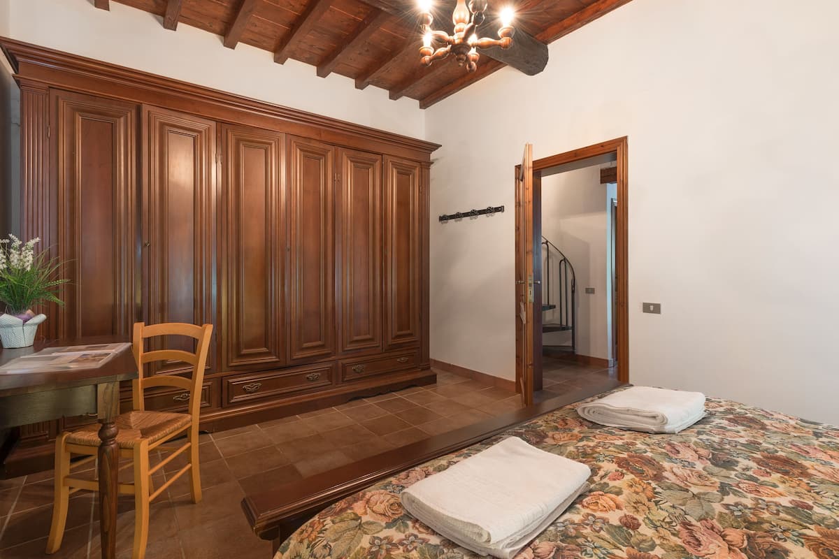 Una habitación con un armario de madera, un escritorio con silla, una cama con edredón estampado y una escalera al fondo.