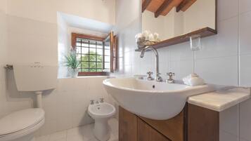 A bathroom with a toilet, bidet, sink, wooden cabinet, and a window with a plant.