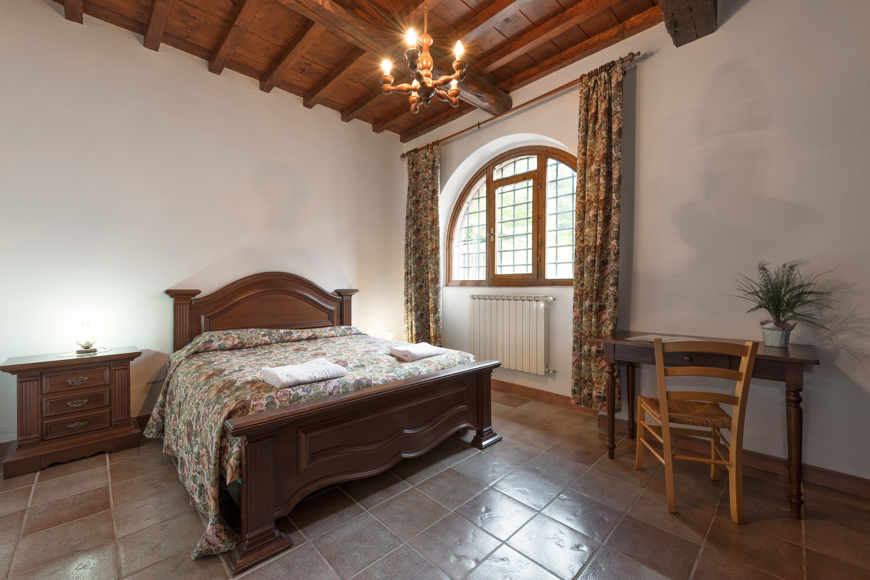 Een slaapkamer met een houten bed, nachtkastje, bureau en stoel. Er is een raam met gordijnen en een radiator.