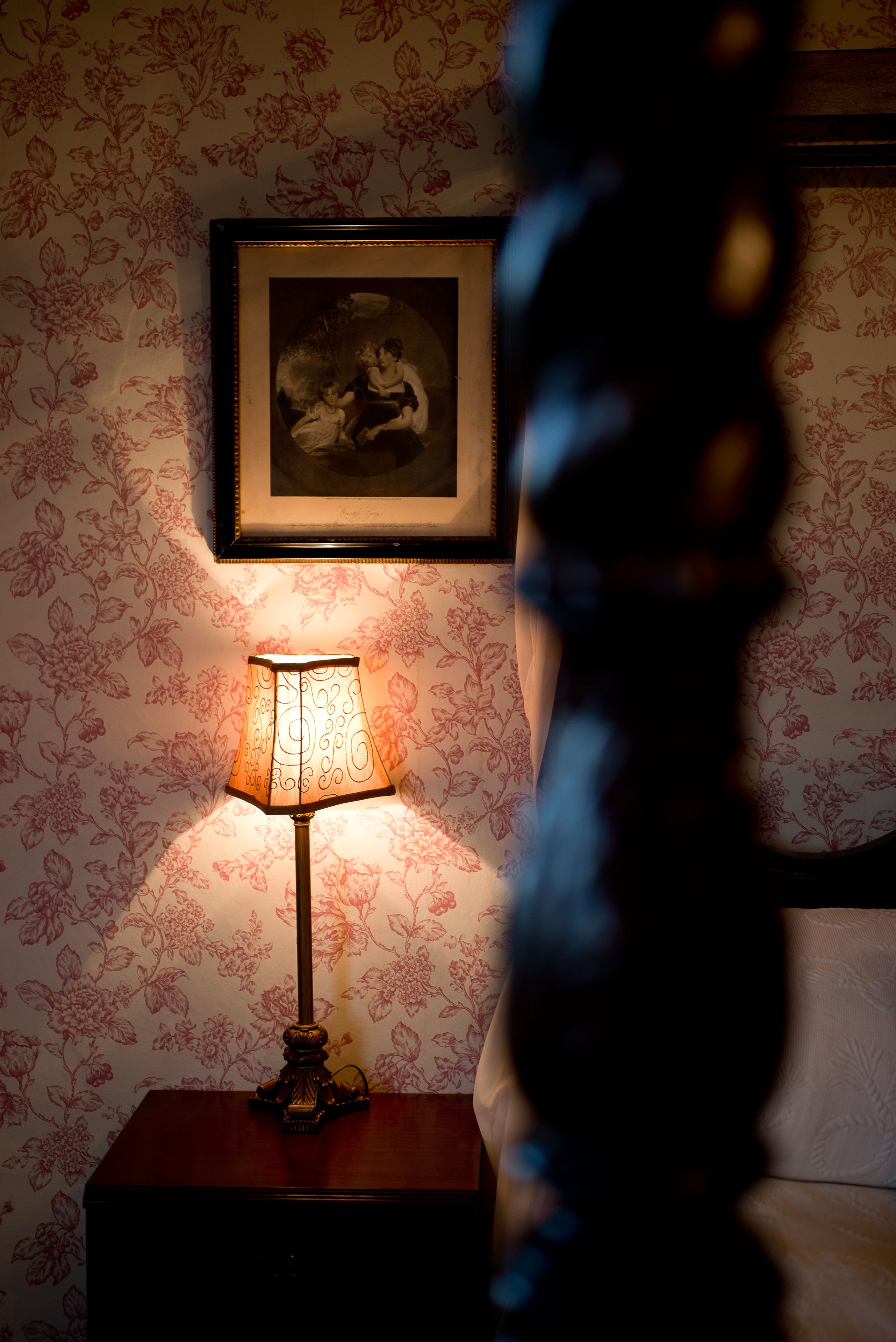 A room with floral wallpaper, a framed picture on the wall, and a table lamp.