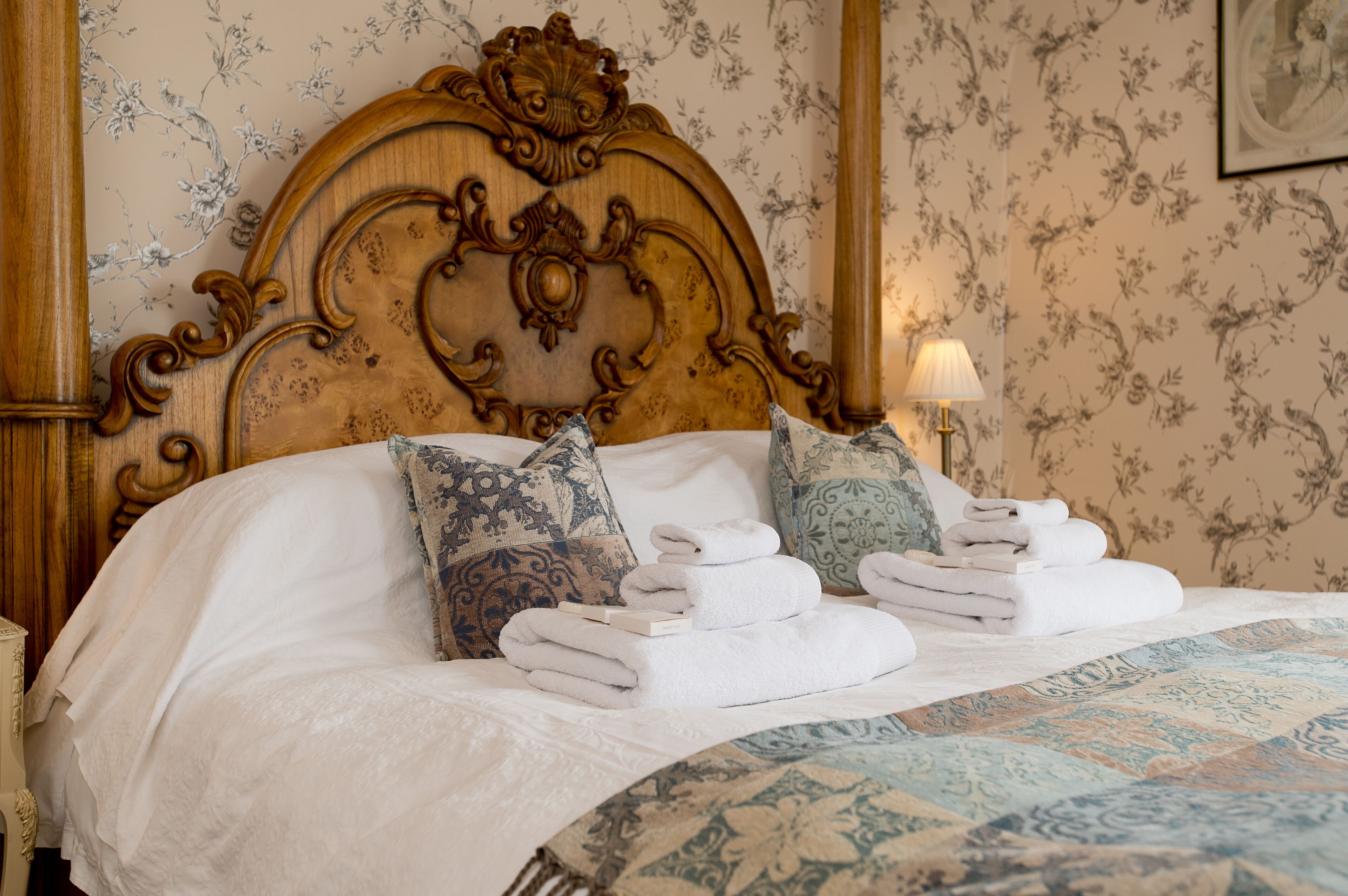 Una cama bien hecha con cabecera de madera, almohadas estampadas y toallas dobladas.