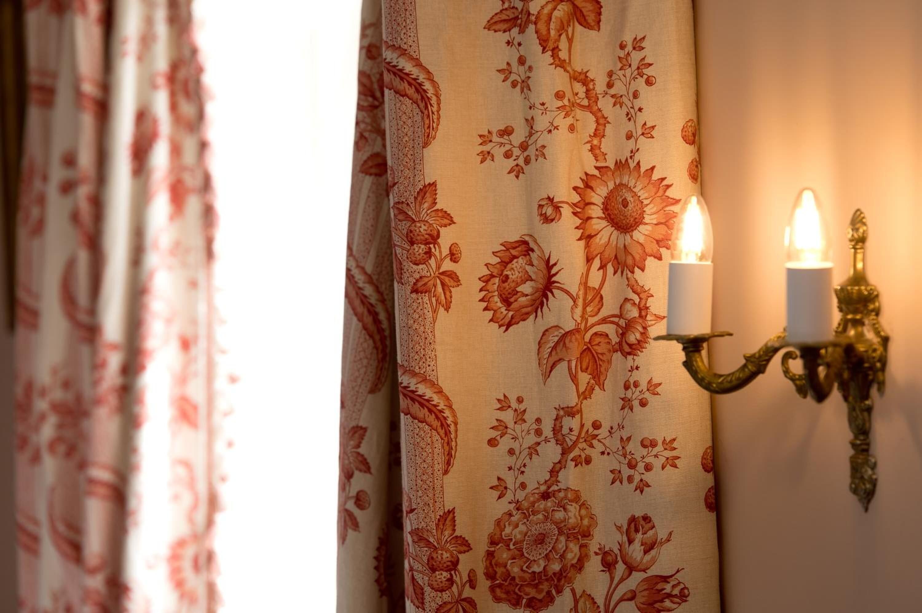 Un candelabro fijado a la pared con dos velas encendidas, una cortina con estampado floral y una ventana con luz natural.