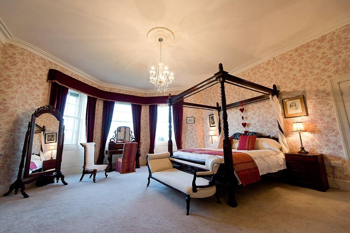 A four-poster bed with a canopy, a sitting chair, a bedside table, a mirror, and a chandelier.