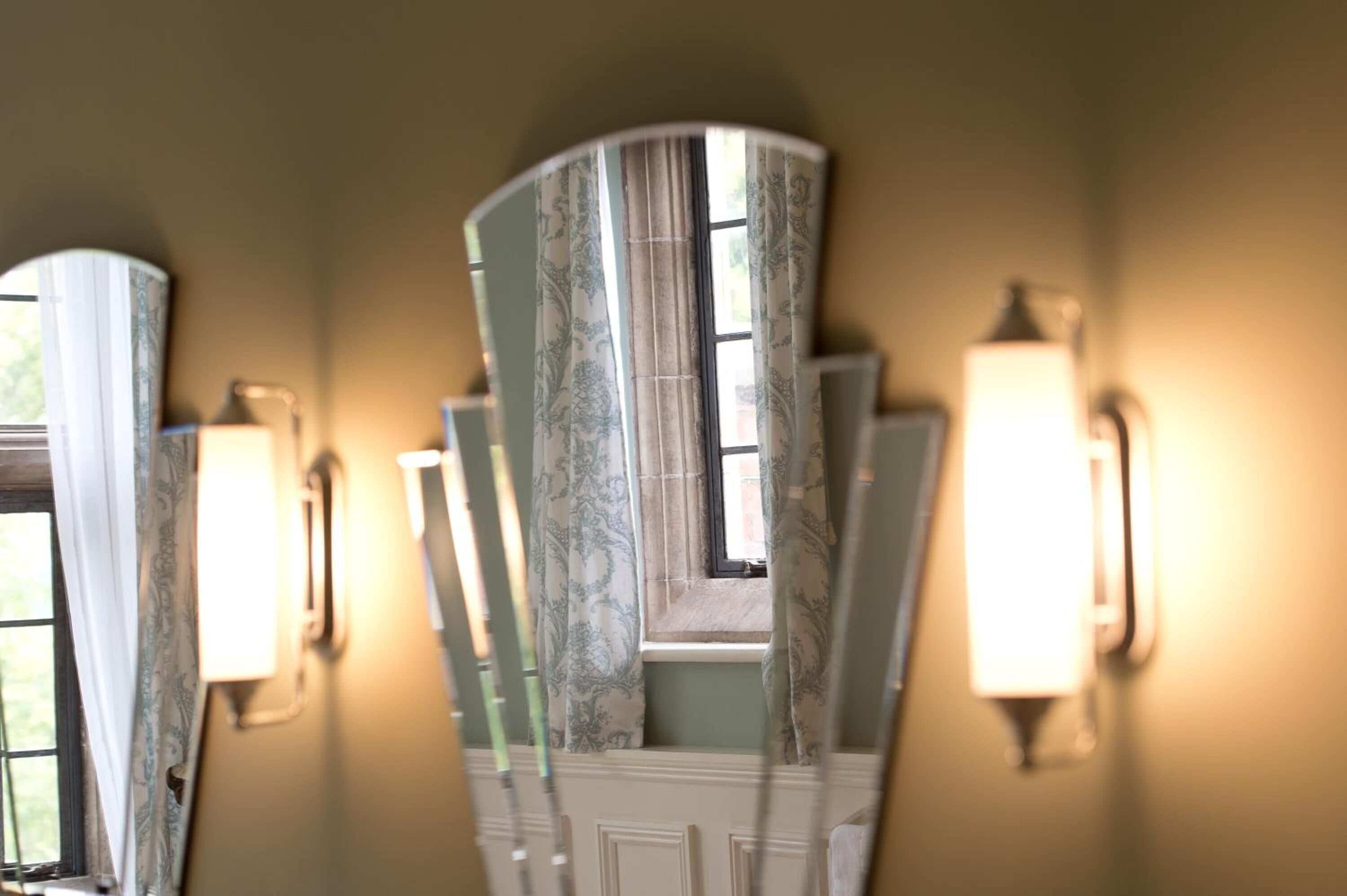 Una habitación con ventana, una lámpara en la pared y un espejo que refleja parte del espacio.
