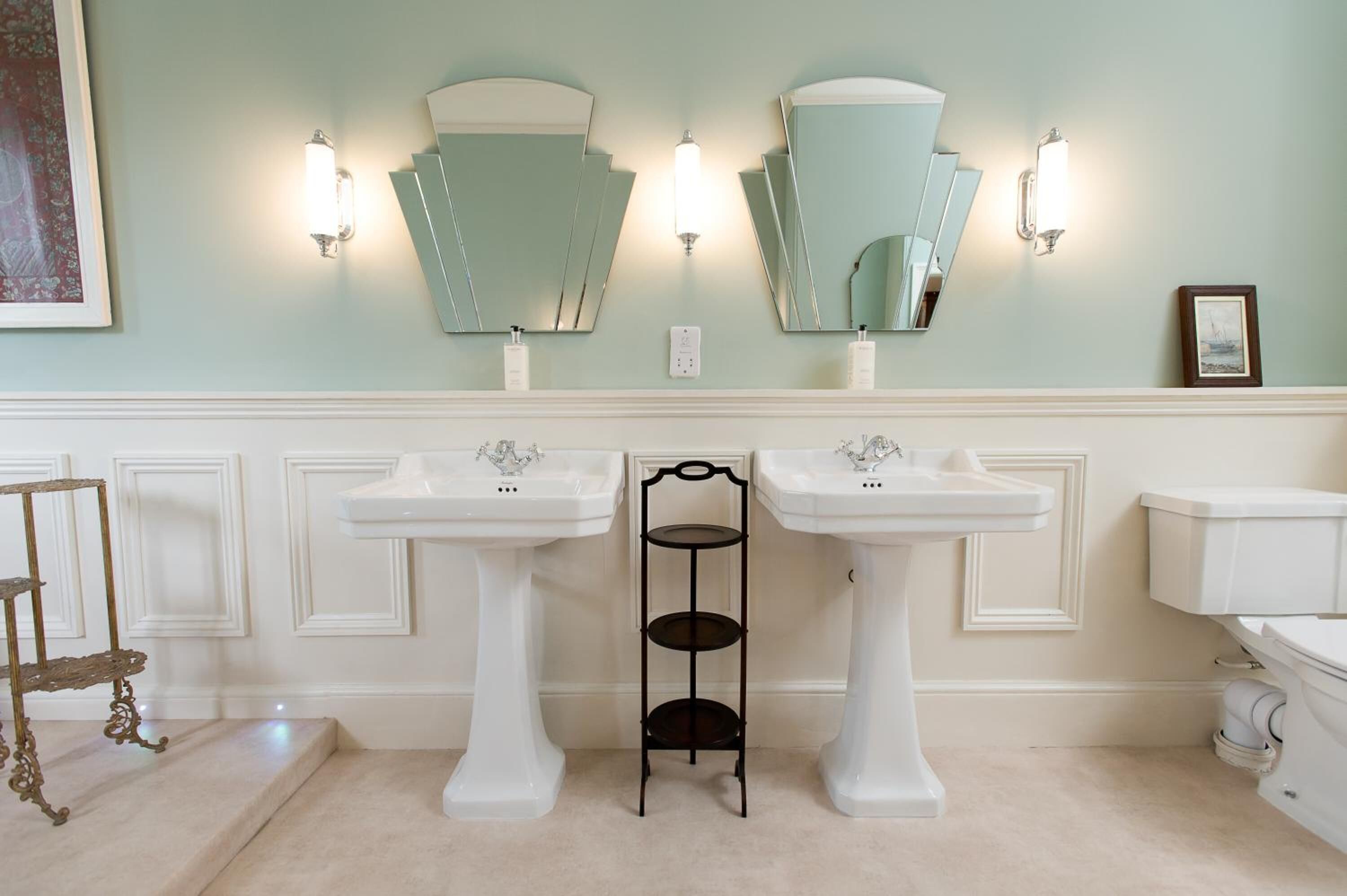 A bathroom with two white pedestal sinks, a black shelf between them, and wall-mounted sconces.