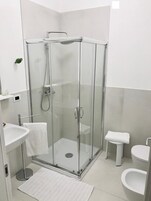 Un baño moderno con ducha envuelta en vidrio, un lavamanos blanco, un inodoro y un taburete blanco pequeño.