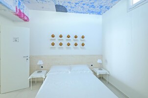 Una habitación de hotel con una cama, dos mesitas de noche, un estante en la pared con objetos decorativos, una puerta con cartel de número de habitación y un techo pintado a medias.