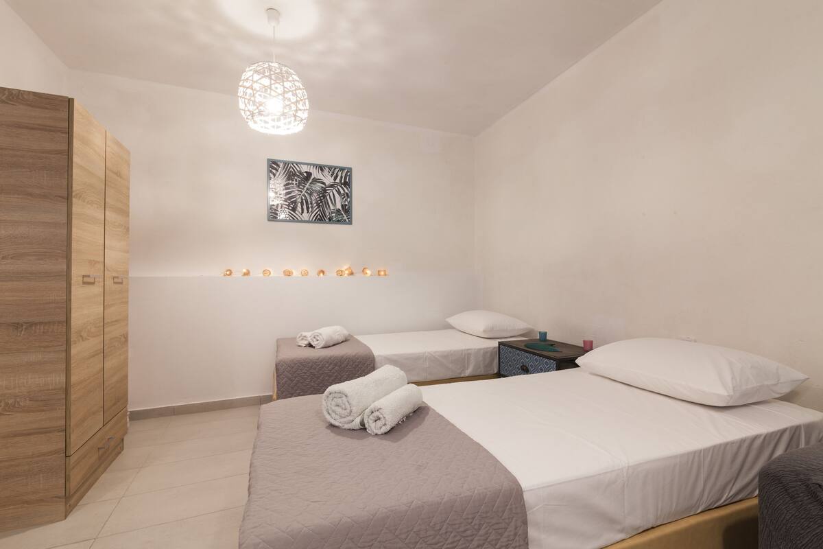 A bedroom with two beds, a wooden wardrobe, a wall-mounted light fixture, and a framed artwork.