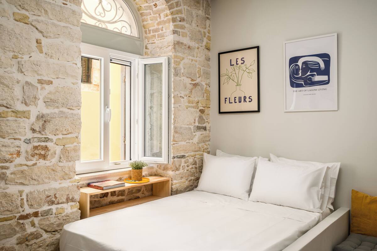 A bedroom with a stone wall, a bed with white bedding, a wooden bedside table with a plant, and two framed artworks.