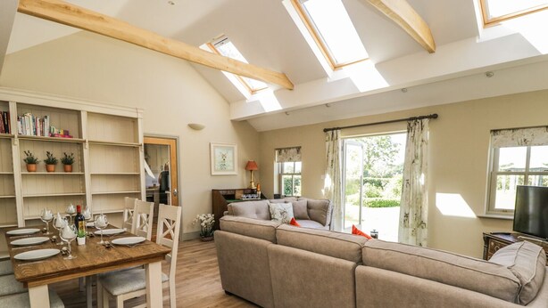 A spacious living area with a wooden dining table set for four, a beige sofa, and a view of the outdoors through large windows. - Providence Cottage (York)
