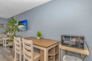 A room with a wooden dining table, two chairs, a small potted plant, a flat-screen TV mounted on the wall, and a microwave on a countertop.