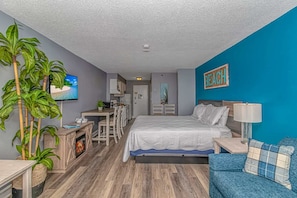 A compact hotel room with a bed, a desk, a kitchenette, and a TV.